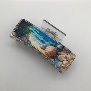 May include: A clear plastic hair clip with a beach scene design. The design features a blue ocean, white sand, palm trees, and seashells.
