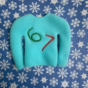 May include: A turquoise felt sweater decoration with the numbers "6" and "7" stitched in green and red thread. The ornament is set against a blue backdrop with white snowflake designs.