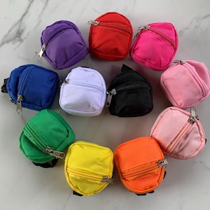 Może przedstawiać: Kolekcja małych, kolorowych plecaków w różnych odcieniach, w tym fioletowym, czerwonym, różowym, niebieskim, białym, czarnym, zielonym, żółtym i pomarańczowym. Każdy miniaturowy plecak posiada zapięcie na zamek błyskawiczny i mały uchwyt.