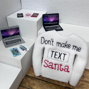 May include: A white long-sleeve sweater with black embroidered text reading Don't make me, a black embroidered box containing the word TEXT, and the word Santa in red embroidery. Miniature laptops and smartphones are displayed nearby.