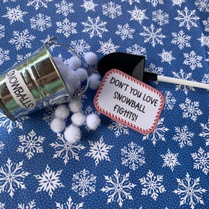 Peut inclure: Un seau en métal argenté avec l'inscription "SNOWBALLS" est renversé, déversant des pompons blancs sur une surface bleue à motifs de flocons de neige. Une petite pelle noire et blanche et une pancarte avec le texte "DON'T YOU LOVE SNOWBALL FIGHTS!" sont également présents.