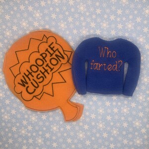 May include: A blue fleece sweater with the text "Who farted?" embroidered on the back. Next to it is an orange whoopie cushion with the text "Whoopie Cushion" embroidered on it.