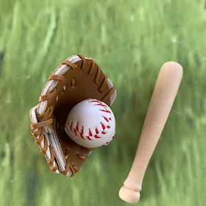 May include: A baseball glove, baseball, and wooden bat are arranged on a green surface. The baseball is white with red stitching, nestled in a brown glove. A light-colored wooden bat rests beside the glove.
