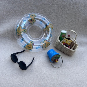 Könnte beinhalten: Ein Miniatur-Set bestehend aus einem transparenten aufblasbaren Schwimmring mit gelben Entenmotiven, einer schwarzen runden Sonnenbrille, einem blauen Plastikeimer mit Sand und einem kleinen roten Apfel sowie einem geflochtenen Korb mit Miniatur-Lebensmitteln.