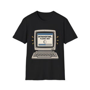 May include: A black short-sleeve t-shirt featuring a graphic of a vintage computer monitor and keyboard. The screen displays a loading bar and the text "INTROVERTING... PLEASE WAIT" in black capital letters against a light background.