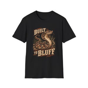Könnte beinhalten: Ein schwarzes Kurzarm-T-Shirt mit einer grafischen Illustration einer geringelten braunen und beigen Schlange. Der Text "BUILT TO BLUFF" ist in einer fetten, hellen Schrift über und unter der Schlange gedruckt, die von Grasbüscheln umgeben ist.