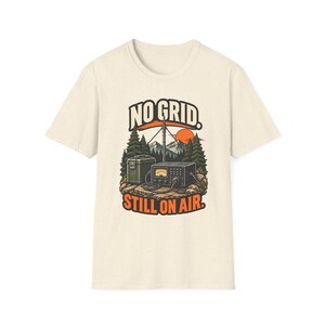 Könnte beinhalten: Ein cremefarbenes Kurzarm-T-Shirt mit einem Grafikdruck, der ein Funkgerät, eine Antenne und eine grüne Aufbewahrungsbox in einer Waldlandschaft mit Bergen und Sonne zeigt. Der Text lautet NO GRID. STILL ON AIR. in fetter Schrift.