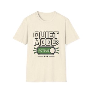 Könnte beinhalten: Ein cremefarbenes Kurzarm-T-Shirt mit dem Text "QUIET MODE:" in fetter, dunkler Schrift über einer Grafik eines Kippschalters. Der Schalter ist grün, enthält das Wort "ACTIVE" und ist von kleinen dekorativen Linien und Punkten umgeben.