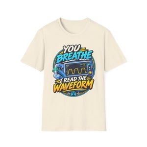 May include: A cream-colored short-sleeve t-shirt featuring a graphic of a medical monitor displaying a waveform. The text reads "YOU BREATHE I READ THE WAVEFORM" in blue and yellow lettering, set against a circular blue and yellow background.