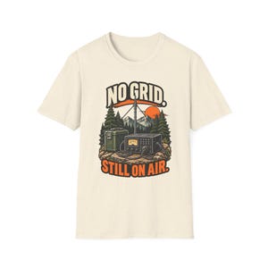 以下が含まれることがあります： クリーム色の半袖Tシャツ。山と太陽がある森の中で、無線機、アンテナ、緑色の収納ボックスが描かれたグラフィックプリントが施されています。太字で「NO GRID. STILL ON AIR.」というテキストが記されています。
