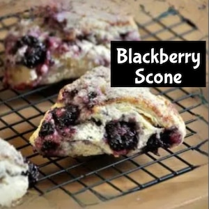 Puede incluir: Primer plano de un bizcocho de mora en una rejilla. El bizcocho es de color marrón dorado y tiene una textura hojaldrada. Está cubierto con moras y una pizca de azúcar en polvo.