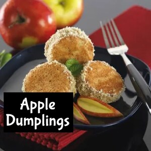 Puede incluir: Tres buñuelos de manzana, espolvoreados con azúcar y canela, están dispuestos en un plato negro con dos rodajas de manzana. Los buñuelos son de color marrón dorado y tienen un exterior crujiente. El plato está sobre un mantel individual rojo.