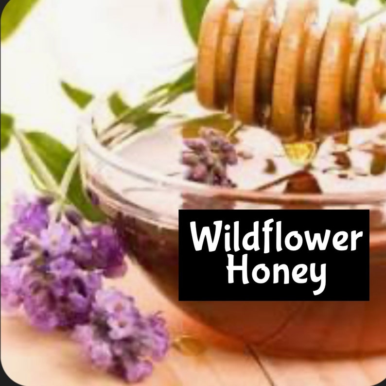 May include: A glass jar filled with golden brown wildflower honey, with a wooden honey dipper and lavender flowers in the background. The jar has a black label that reads "Wildflower Honey".