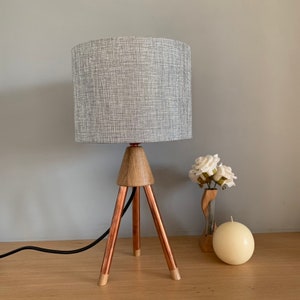 May include: A stylish table lamp with a grey fabric shade. The lamp features a wooden base and three copper legs. A black power cord is attached. A vase with white flowers and a spherical candle are next to the lamp.