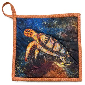 May include: A quilted potholder featuring a sea turtle design. The turtle is primarily orange and brown, set against a dark blue and black background with gold accents. The potholder has an orange border and a loop for hanging.