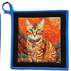 May include: Square quilted pot holder featuring a detailed illustration of a tabby cat with green eyes and a colorful beaded collar. The cat is set against an orange and patterned background, framed by black and blue fabric. Includes a blue hanging loop.
