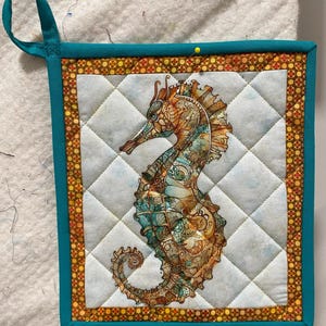 May include: Quilted seahorse pot holder with a teal border and hanging loop. The seahorse is detailed with shades of teal, orange, and brown. The pot holder has a diamond pattern and a decorative border with orange and yellow dots.