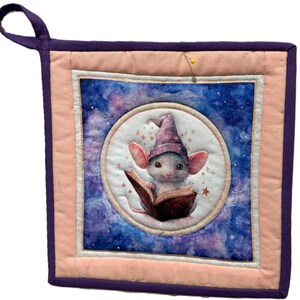 Puede incluir: Un salvamanteles cuadrado acolchado con un diseño caprichoso. El centro presenta un ratón blanco con un sombrero de mago morado, leyendo un libro sobre un fondo estrellado. El salvamanteles tiene un centro color melocotón, un borde blanco y un ribete morado.