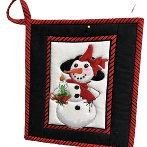 May include: A square, quilted pot holder featuring a snowman design. The snowman has a carrot nose, black eyes, and a red scarf and bow. The pot holder has a black background with a red and black striped border and a red loop for hanging.