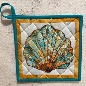 May include: A teal and gold quilted hot pad with a detailed seashell design. The seashell is a light blue, brown, and gold color.
