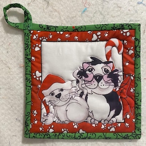 Könnte beinhalten: Ein roter und grüner gesteppter Topflappen mit einer weißen Mitte, auf der zwei Katzen abgebildet sind. Eine Katze trägt einen Weihnachtsmann-Hut und die andere Katze hält einen Zuckerstock. Der Rand des Topflappens hat ein grünes und weißes Wirbelmuster mit Pfotenabdrücken.