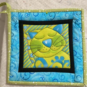 Puede incluir: Un posavasos acolchado verde y azul con un diseño de gato de dibujos animados. El gato tiene los ojos cerrados y una gran sonrisa. El posavasos está rodeado por un borde azul y verde con un patrón de espiral.