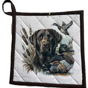 May include: Square quilted pot holder with a brown border and hanging loop. The centre features a detailed illustration of a brown Labrador Retriever, ducks, and bulrushes. The background is white with a diamond pattern.