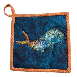 May include: Square quilted potholder with a whale design. The whale is a mix of blue, yellow, and orange, set against a dark blue background with a floral pattern. The potholder has an orange border and a loop for hanging.