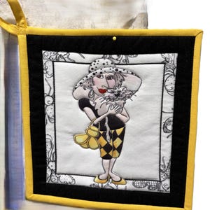 May include: A square quilted potholder with a bright yellow border and a black frame. The center features an embroidered illustration of a person in a polka-dot hat holding a cat, wearing a black top and yellow and black diamond-patterned pants.