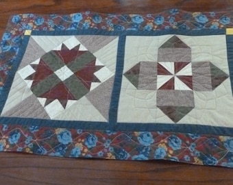 Könnte beinhalten: Ein gesteppter Tischläufer mit zwei verschiedenen Patchwork-Designs. Der Läufer hat einen blauen, roten und grünen floralen Rand.