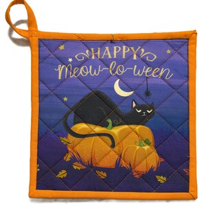 Puede incluir: Manopla acolchada con temática de Halloween, con borde y lazo para colgar de color naranja. El diseño presenta un gato negro sentado sobre calabazas bajo una luna, con las palabras "HAPPY Meow-lo-ween". El fondo es azul oscuro con estrellas y una araña.