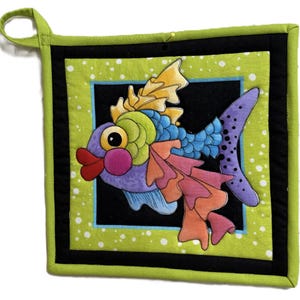 May include: A colorful, quilted potholder featuring a whimsical fish design. The fish has a purple body, blue scales, and orange and pink fins. The potholder has a black border, a lime green background with white polka dots, and a green loop for hanging.