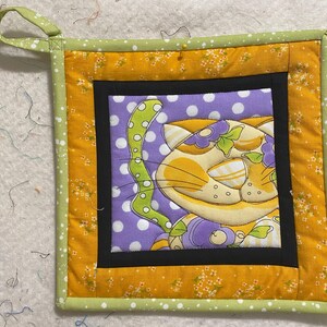 Peut inclure: Un dessous de plat matelassé jaune et vert avec un motif de chat violet et blanc. Le chat porte un nœud vert et est entouré de pois blancs.