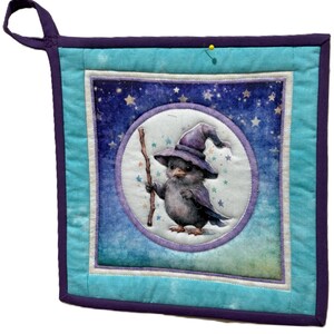 May include: A square, quilted potholder with a whimsical design. The center features a cartoon bird wearing a wizard hat and holding a staff, set against a starry night sky. The potholder has a teal and purple color scheme with a hanging loop.