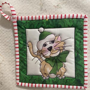 Könnte beinhalten: Ein grüner und weißer gequilteter Topflappen mit einem roten und weißen Tupfenrand. In der Mitte befindet sich eine Comic-Katze, die einen grün-weißen Weihnachtsmann-Hut mit einer grünen Schleife trägt.