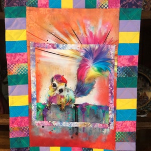 May include: A colorful quilt with a framed image of a squirrel with a rainbow tail. The quilt is made of various fabrics with a patchwork design. The colors include pink, yellow, blue, green, and purple.