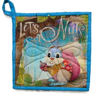 Könnte beinhalten: Quadratischer, gesteppter Topflappen mit blauem Rand und Aufhängeschlaufe. In der Mitte befindet sich ein Eichhörnchen, das eine Eichel hält, mit dem Text "Let's Get Nuts" in einer verspielten Schriftart. Der Hintergrund hat ein Baum- und Blättermuster.