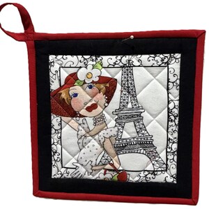 May include: Quilted pot holder with a red border and hanging loop. The center features a cartoon woman in a red hat and the Eiffel Tower. The design is framed by a black border and white quilted background.