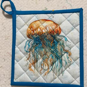 May include: A blue and white quilted hot pad with a colorful jellyfish illustration. The hot pad has a blue fabric border and a loop handle.