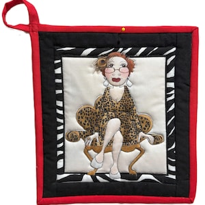 May include: A quilted pot holder featuring a cartoon woman in a leopard print dress seated in a gold chair. The pot holder has a red border, black and white zebra print, and a black inner border. A loop is attached for hanging.