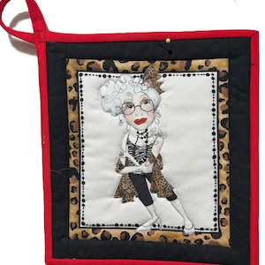May include: A quilted hot pad featuring a whimsical illustration of a stylish woman with glasses, a zebra-striped top, and a leopard-print shawl. The pad has a red border, black backing, and a leopard print accent.