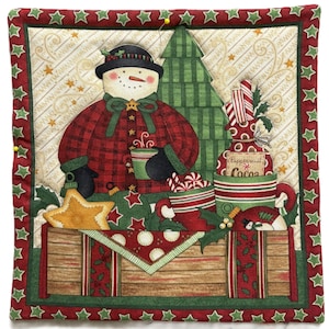 Puede incluir: Un edredón cuadrado con temática navideña con un muñeco de nieve con abrigo a cuadros, un árbol de Navidad verde y regalos. El edredón tiene un borde de estrellas rojas y verdes e incluye el texto "Peppermint Cocoa".