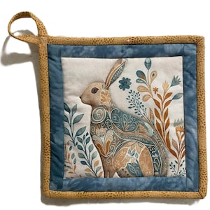 May include: A brown and beige patterned rabbit with blue and brown floral accents is featured on a quilted pot holder with a brown border and a blue inner border.