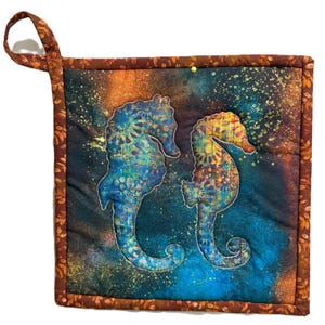 May include: A quilted potholder featuring two seahorse silhouettes in shades of blue and yellow, set against a dark blue and orange background with gold speckles. The potholder has a brown floral border and a matching loop for hanging.