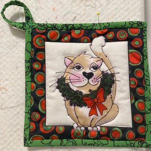 Könnte beinhalten: Ein grüner und schwarzer gesteppter Topflappen mit einer weißen Mitte, auf der ein Comic-Katze mit einem grünen Kranz und einer roten Schleife zu sehen ist. Die Katze hat rosa Wangen und einen weißen Fleck auf der Brust.