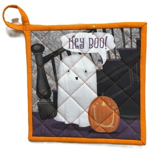 Pode incluir: Um pegador de panela acolchoado com tema de Halloween com borda e laço laranja. O design apresenta um fantasma branco dizendo "Hey BOO!" ao lado de uma abóbora laranja. O fundo mostra uma escada.