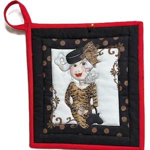 May include: A square, quilted hot pad with a red border and hanging loop. The center features a whimsical illustration of a woman in a tiger-striped dress, a black hat, and a leopard print bow. The background is white with brown floral accents.