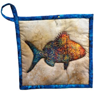 May include: A square, quilted potholder featuring a colorful fish applique. The fish has a blue tail and a body with orange, yellow, and blue patterns. The potholder has a blue border and a loop for hanging. A pin is holding the layers together.