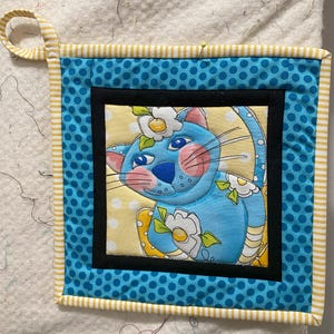 Peut inclure: Un dessous de plat matelassé avec un chat bleu orné de fleurs blanches sur un fond jaune à pois. Bordure bleue à pois, reliure rayée jaune et blanche. Une boucle est attachée pour suspendre.