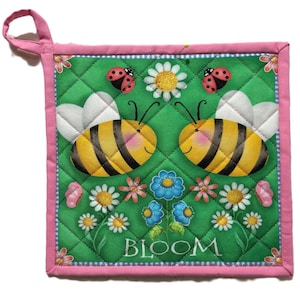 Peut inclure: Un dessous de plat matelassé avec un fond vert représentant deux abeilles de dessin animé, des coccinelles et des fleurs. Le mot "BLOOM" est imprimé en bas. Le dessous de plat a une bordure rose et une boucle rose pour l'accrocher.
